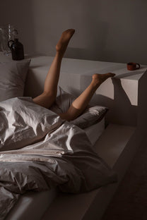 Person lying in bed with legs extended over a nightstand in a dimly lit room.